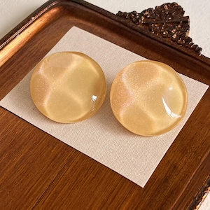 May include: A pair of round, translucent, pale yellow stud earrings with a subtle, shimmering, cat-eye effect. The earrings have a smooth, domed surface that reflects light, creating a soft, pearlescent glow across the circular, polished resin material.