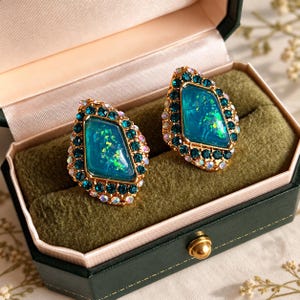 May include: A pair of gold-tone earrings featuring a central teal stone with iridescent flecks, surrounded by a border of dark teal crystals and an outer edge of clear, shimmering rhinestones, presented in a dark green velvet box.