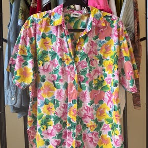 May include: A short-sleeved button-down shirt featuring a vibrant floral pattern in shades of pink, yellow, and green. The garment includes a classic collar, a single chest pocket on the left side, and white buttons running down the center front.