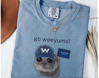 May include: A blue jean colored t-shirt featuring a graphic of a small animal wearing a blue cap with a white W logo. The text go weeyums! is printed above the animal, and a small blue flag with the text WILLIAMS RACING is visible nearby.