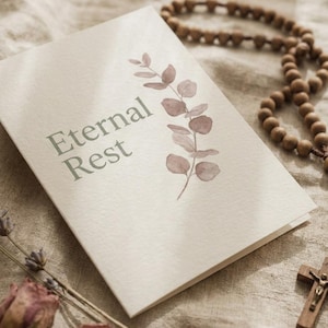Pode incluir: Um cartão dobrado de cor creme com o texto Eternal Rest numa fonte serifada verde-escura, acompanhado por uma ilustração em aguarela de um ramo de eucalipto em tons malva suaves, colocado junto a contas de oração de madeira e um crucifixo.