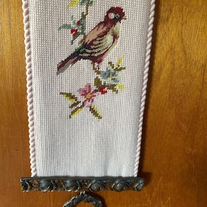 May include: A vertical needlepoint wall hanging on a pale pink fabric background featuring a brown and red bird perched on a branch with blue and pink flowers. The piece is framed by a twisted rope border and a dark metal base with floral carvings.