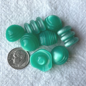 May include: A collection of seven translucent mint green buttons in various shapes, including domed, ribbed, and circular designs, displayed next to a silver United States dime for scale, showing the relative size of these small sewing fasteners.