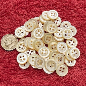 May include: A collection of off-white, four-hole buttons scattered on a red textured surface. A U.S. dime is included for size comparison. The buttons have a pearlescent sheen and are likely made of mother-of-pearl.