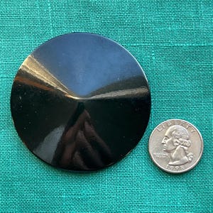 May include: A large, circular, black button with a smooth, glossy finish and a slightly raised, conical center. The button is positioned next to a standard United States quarter coin for scale, which measures approximately 0.955 inches in diameter.