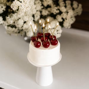 May include: A small round cake with smooth white frosting, topped with several dark red cherry-shaped candles. Each candle features a thin gold stem and a lit flame. The cake sits on a simple white pedestal stand, creating a clean and bright display.