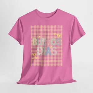 May include: A pink t-shirt featuring the phrase "BUENOS DIAS" in colorful, patchwork-style letters. The text is set against a pink and tan plaid background, with yellow butterfly accents.