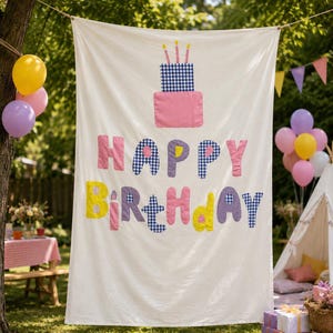 May include: A white fabric banner featuring a two-tiered birthday cake graphic with three candles at the top. The text HAPPY BIRTHDAY is appliqued in a mix of pink, yellow, purple, and blue-and-white checkered fabric letters across the center.