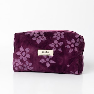 May include: A rectangular quilted pouch in deep purple with a lighter purple floral pattern. The bag features a pink zipper closure and a central cream-colored fabric label with the text Jelita Handmade in Indonesia printed in black ink.