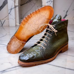 May include: An olive green ankle boot with a textured crocodile-embossed leather upper, cream-colored laces, and silver eyelets. The boot features a translucent amber-toned rubber lug sole with a wavy tread pattern and a pink interior lining.