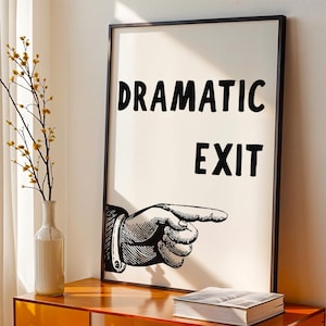 May include: A framed art print with the words "DRAMATIC EXIT" and a pointing hand illustration. The print is displayed on an orange cabinet with books and a vase with branches. A yellow chair and white curtains are in the background.