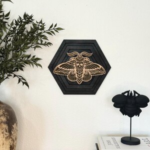 May include: A black wooden wall art piece in a hexagonal shape, featuring a detailed moth design with a skull on its head. The moth is light brown, set against a black background. A black sculpture of a bee is on a stand, and books are stacked below.