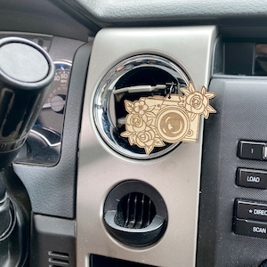 May include: A wooden car air freshener shaped like a camera, adorned with floral accents. The camera design features a lens and is surrounded by roses and other flowers. The air freshener hangs from a black string and is set against a car's dashboard.