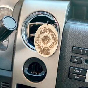 May include: Oval-shaped wooden car air freshener with a laser-etched design of a crystal ball, an eye, and hands. The air freshener is hanging from a car vent. The design is in a light brown color.