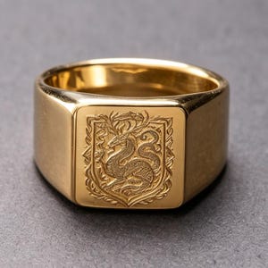 May include: A gold-toned signet ring featuring a flat, square face with an intricate, laser-engraved design of a mythical dragon creature centred within a decorative shield-like border. The band is smooth, polished, and has a wide, solid construction.