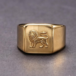 May include: A gold-toned signet ring featuring a flat, rectangular face with an embossed lion figure in the center. The lion is depicted in profile, standing with its tail curved upward. The ring has a polished, smooth finish throughout the band.