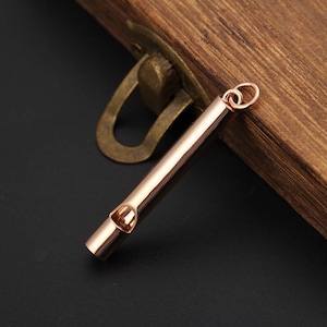 May include: A rose gold-toned cylindrical whistle pendant featuring a smooth, polished metallic finish. The whistle has a small circular opening on the side and includes a small jump ring attached to the top end for hanging or attachment purposes.