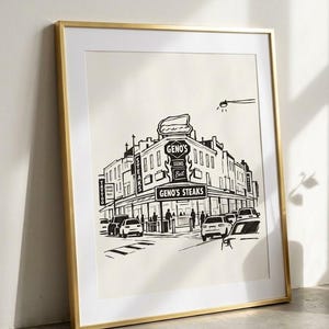 Könnte beinhalten: Ein gerahmter schwarz-weißer Strichzeichnungsdruck, der das Restaurant Geno's Steaks an einer Straßenecke zeigt. Die Illustration enthält Autos, Straßenschilder und den Text Geno's Steaks, Steaks und Tattoo. Der Druck ist in einem Goldrahmen.