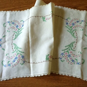 May include: Cream-colored embroidered table runner with a scalloped edge. The runner features floral embroidery in pink, blue, and green. The design includes a central floral motif and a decorative border.