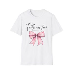 Puede incluir: Una camiseta blanca de manga corta con un gráfico de un lazo rosa centrado en el pecho. Sobre el lazo, el texto Faith over fear aparece en una fuente cursiva negra, con el texto 2 TIMOTHY 1:7 impreso directamente debajo.
