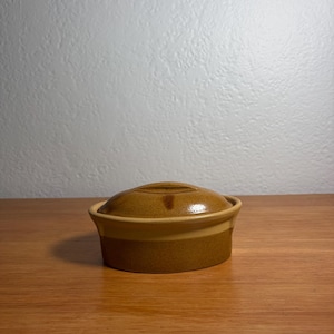 May include: A small, oval-shaped ceramic casserole dish with a matching lid. The piece features a speckled, earthy brown glaze with a lighter tan band encircling the upper portion of the base. The lid has a simple, raised handle in the center.