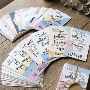 May include: A collection of rectangular cards featuring various religious quotes and scripture verses in black cursive and print text. The cards have abstract pastel-coloured backgrounds in shades of blue, pink, and yellow, and one stack is tied with twine.