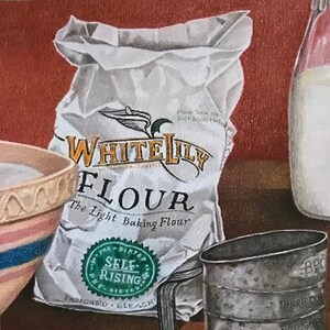 May include: A white bag of White Lily Flour, the light baking flour, with a green self-rising seal. The bag is sitting on a wooden surface next to a white pitcher and a metal measuring cup.