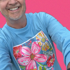 May include: Light blue long-sleeve shirt with a floral design. The graphic features large pink, red, and yellow flowers with green and blue accents. The word "bloom" is printed in pink at the bottom of the design.