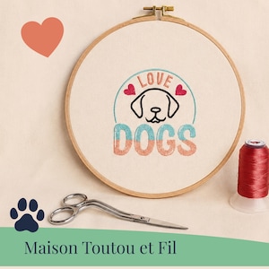 Peut inclure: Un tambour à broder rond en bois contenant un tissu blanc avec un motif brodé représentant une tête de chien, deux cœurs rouges et le texte LOVE DOGS en bleu et orange. À côté se trouvent des ciseaux en métal argenté et du fil rouge.