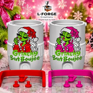 Könnte beinhalten: Zwei weiße zylindrische Getränkekühler mit einer Grafik einer grünen Figur mit Weihnachtsmütze und dem Text Grinchy But Boujee. Ein Kühler hat rote Akzente und einen roten Griff, der andere hat pinke Akzente und einen pinken Griff.