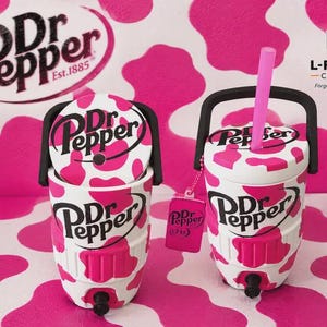 Pu&ograve; includere: Due refrigeratori per bevande Dr Pepper con stampa a macchie di mucca bianche e rosa, manici neri e rubinetti. Uno ha una cannuccia rosa e un'etichetta rosa Dr Pepper attaccata. Entrambi gli articoli mostrano il logo Dr Pepper in nero.