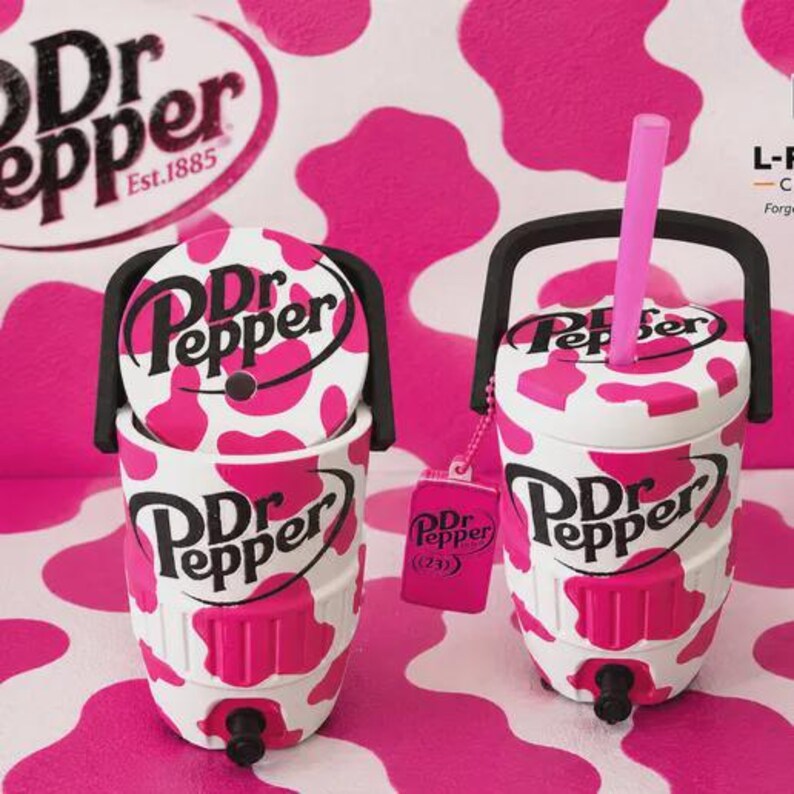 Pu&ograve; includere: Due refrigeratori per bevande Dr Pepper con stampa a macchie di mucca bianche e rosa, manici neri e rubinetti. Uno ha una cannuccia rosa e un'etichetta rosa Dr Pepper attaccata. Entrambi gli articoli mostrano il logo Dr Pepper in nero.