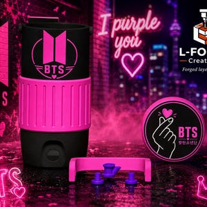 May include: A black cylindrical container featuring a bright pink BTS logo and a matching pink textured grip band. Beside it sits a circular black lid with a white graphic of a finger heart and the text BTS. A small pink bracket with three purple pegs.