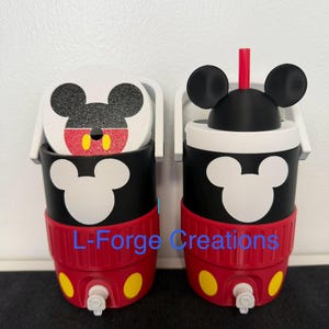 Può includere: Due refrigeratori per bevande neri e rossi con manici e rubinetti bianchi. I refrigeratori presentano una silhouette bianca di Topolino sul davanti, cerchi gialli sulla base e un coperchio con orecchie nere di Topolino e una cannuccia rossa. Testo: L-Forge Creations.