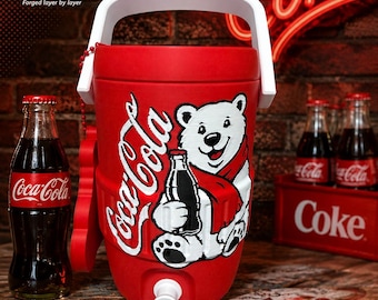 Può includere: Un refrigeratore per bevande cilindrico rosso con manico bianco e una grafica di un orso polare bianco con una sciarpa rossa che tiene in mano una bottiglia di Coca-Cola. Il refrigeratore ha un rubinetto bianco alla base e il logo Coca-Cola.