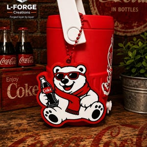 May include: A flat, die-cut plastic ornament featuring a white polar bear wearing red sunglasses and a red scarf, holding a black glass bottle with a red Coke label. The ornament hangs from a red beaded chain against a red plastic cooler.