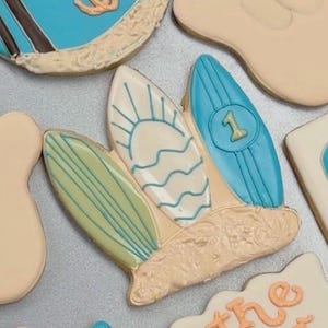 Puede incluir: Galletas de azúcar decoradas con temática playera. Los diseños incluyen tablas de surf, una palmera, chanclas y un gesto con la mano. Algunas galletas tienen el texto "one" y "the big one" en glaseado naranja. Las galletas están en una bandeja plateada.