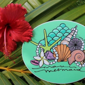 May include: A red hibiscus flower and a green oval sticker with a seashell design. The sticker features a starfish, pearls, and the words "maui mermaid" in a script font. The background is a green palm leaf.