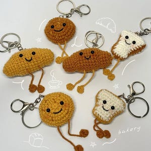 May include: A collection of six crocheted keychains featuring various baked goods including round cookies, croissants, and slices of toast. Each item has a smiling face with black bead eyes, dangling yarn legs, and a silver metal keyring attachment.