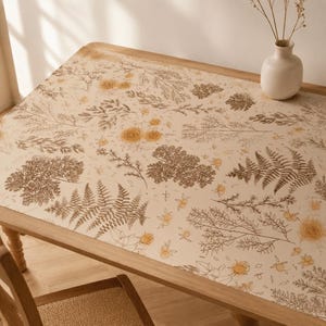 May include: A rectangular table covered with a cream-colored tablecloth featuring a botanical print of brown ferns, thin branches, and yellow flowers. A white ceramic vase containing dried stems sits on the corner of the table.