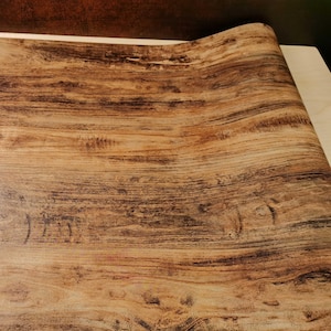 May include: A roll of adhesive contact paper featuring a realistic wood grain pattern. The surface displays a mix of light tan and dark brown tones with horizontal streaks, knots, and natural wood textures printed across the entire material.