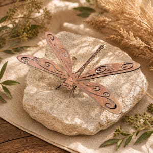 Pode incluir: Uma libélula de metal cor de cobre com padrões intrincados de espirais recortados nas quatro asas e um corpo segmentado. A libélula está posicionada plana sobre uma pedra natural de cor bege claro, texturizada e de forma irregular.