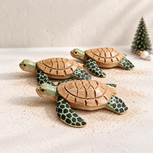 May include: Three hand-carved wooden sea turtle figurines featuring natural light brown shells with carved geometric patterns and light green heads and flippers decorated with dark green spots. Each turtle is positioned in a crawling pose.