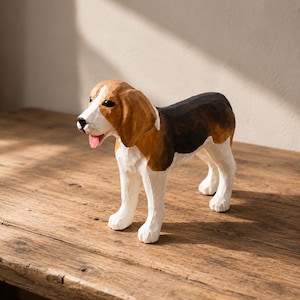 May include: A hand-carved wooden figurine of a beagle dog. The sculpture features a tri-colour coat with white legs and chest, brown ears and face markings, and a black back. The dog has a black nose, dark eyes, and a small pink tongue showing.