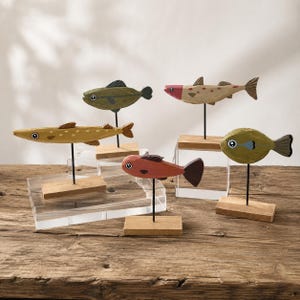 Könnte beinhalten: Fünf kleine, handbemalte Holzfischfiguren in verschiedenen Formen und Farben, darunter Grün, Gelb, Rot und Hellbraun. Jeder Fisch ist auf einem dünnen schwarzen Metallstab befestigt, der auf einem rechteckigen Holzsockel montiert ist.