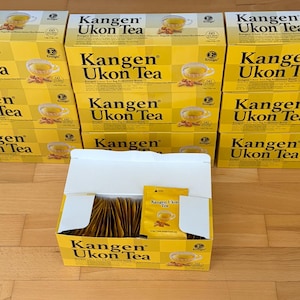 Op de afbeelding: Meerdere gele dozen Kangen Ukon Thee zijn opgestapeld, met één doos open om individuele theezakjes te laten zien. De dozen hebben zwarte tekst en een theekopje-afbeelding. Dit product is waarschijnlijk een gezondheids- en welzijnsthee.