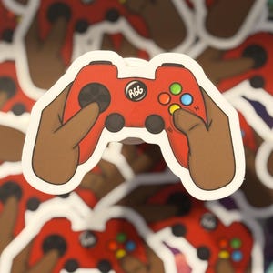 May include: A die-cut sticker featuring a red video game controller held by two brown hands. The controller has black joysticks, a central circular button with the text RGG, and four colored action buttons in green, blue, yellow, and red.