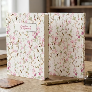 Può includere: Un taccuino con copertina rigida su sfondo bianco caratterizzato da un motivo ripetuto di fiori rosa, foglie verdi, rami marroni e piccoli uccelli. Un'etichetta bianca rettangolare sulla copertina riporta la parola Notebook in corsivo.