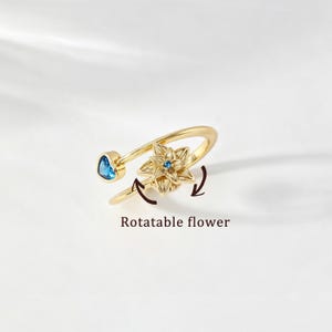 Könnte beinhalten: Ein goldfarbener Ring mit einer drehbaren Blumenverzierung, die einen kleinen blauen Stein in der Mitte hat, sowie einem herzförmigen blauen Stein am Ende der Ringschiene. Der Text Rotatable flower steht unter dem Ring mit zwei Pfeilen.