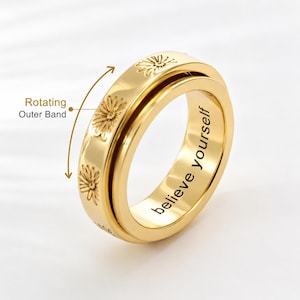 Op de afbeelding: Een goudkleurige metalen spinnerring met een buitenband gegraveerd met madeliefjesmotieven. De binnenzijde van de ring is gegraveerd met de tekst "believe yourself". Een pijlgrafiek geeft de draaiende beweging van de buitenband aan.